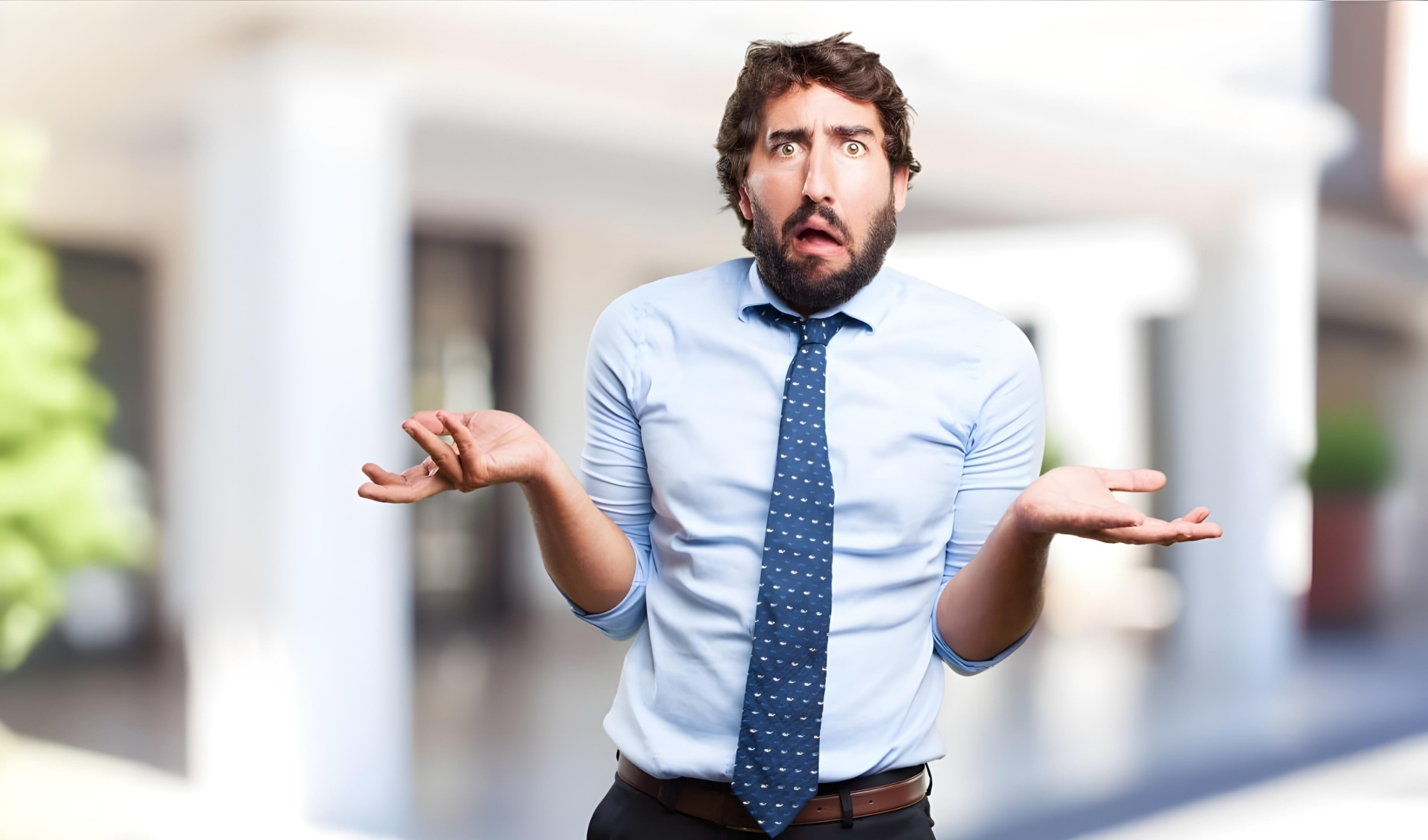 A man with curly hair and a beard stands outdoors wearing a light blue shirt and polka-dot tie. He has an expressive, confused look on his face, raising his eyebrows and shrugging with both hands open. The background is blurred.