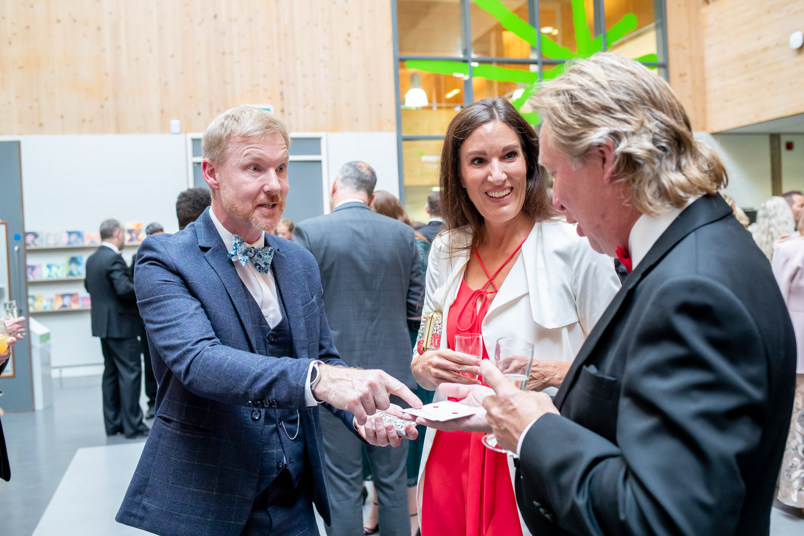 Three people in formal attire are engaged in a conversation at a social event. One man in a blue suit gestures with playing cards, illustrating strategic recruitment planning through magic. A woman in a white outfit and a man in a black suit watch him intently.